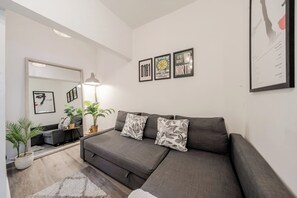 Ein modernes Wohnzimmer mit einem grauen Sofa, zwei dekorativen Kissen, einem kleinen Beistelltisch, einer Stehlampe und einem Spiegel. An der Wand hängen gerahmte Bilder und es gibt zwei Zimmerpflanzen.