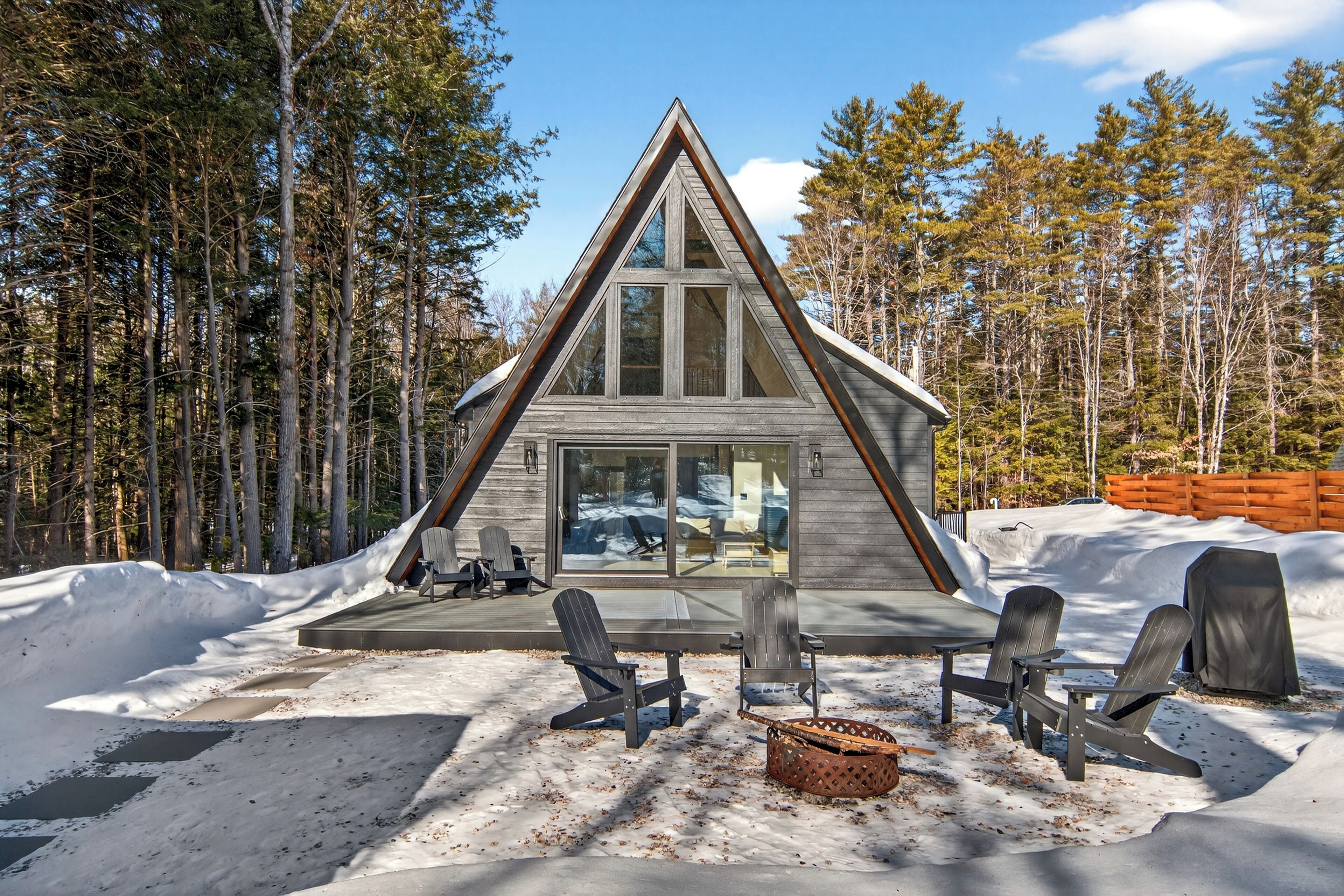 A cabin with a triangular roof, large windows, and a deck with Adirondack chairs.