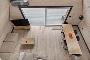 A compact room with a wooden floor, a sofa, a small table, a dining table with chairs, and a large window with blinds. - Modern A-Frame w/Sauna - Near Ragged Mtn, NH (Danbury)