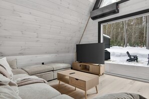 A modern interior with a wooden floor, a sectional sofa, a television on a wooden stand, and a view of a snowy landscape through a large window. - Modern A-Frame w/Sauna - Near Ragged Mtn, NH (Danbury)