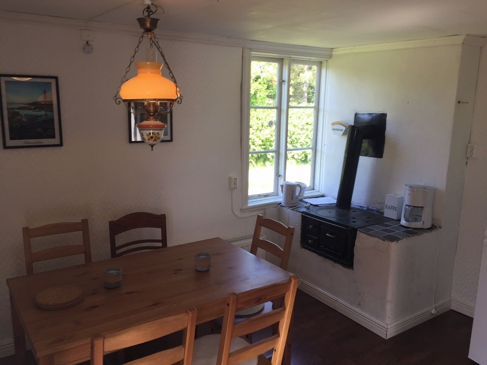 A small kitchen with a wooden table and chairs, a coffee maker, and a window with a view of greenery.