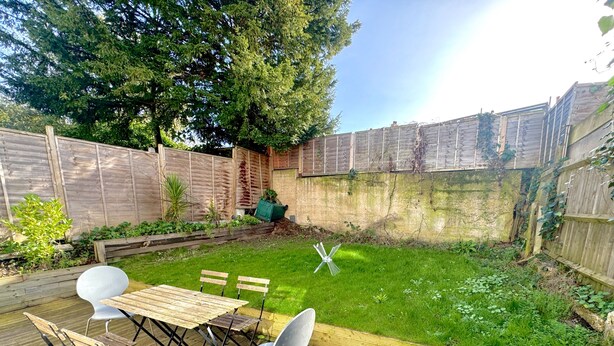 A wooden deck with a table and chairs, surrounded by a grassy backyard with a wooden fence and trees. - North West London 3-Bed Flat, Sleeps 7 with Garden (London)