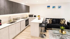 A modern kitchen with a central island, a dining area with a black sofa and a glass coffee table, and a living room with a black couch. - North West London 3-Bed Flat, Sleeps 7 with Garden (London)