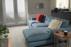 Ett vardagsrum med en blå soffa, en röd kudde, en mönstrad kudde, en golvlampa, ett litet runt bord med en kopp på och en krukväxt.
