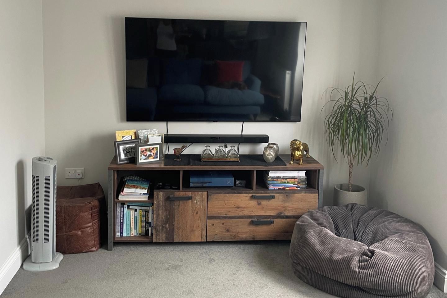 Een woonkamer met een flatscreen televisie aan de muur, een houten dressoir met boeken en decoratieve objecten, een bruin leren poef en een potplant.