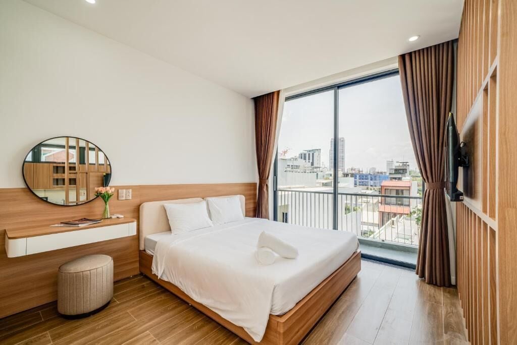 A hotel room with a large bed, a round mirror, a small table, and a view of the city through a large window.