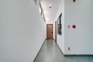 Corredor com porta de madeira no final, iluminação embutida e um detector de incêndio na parede.