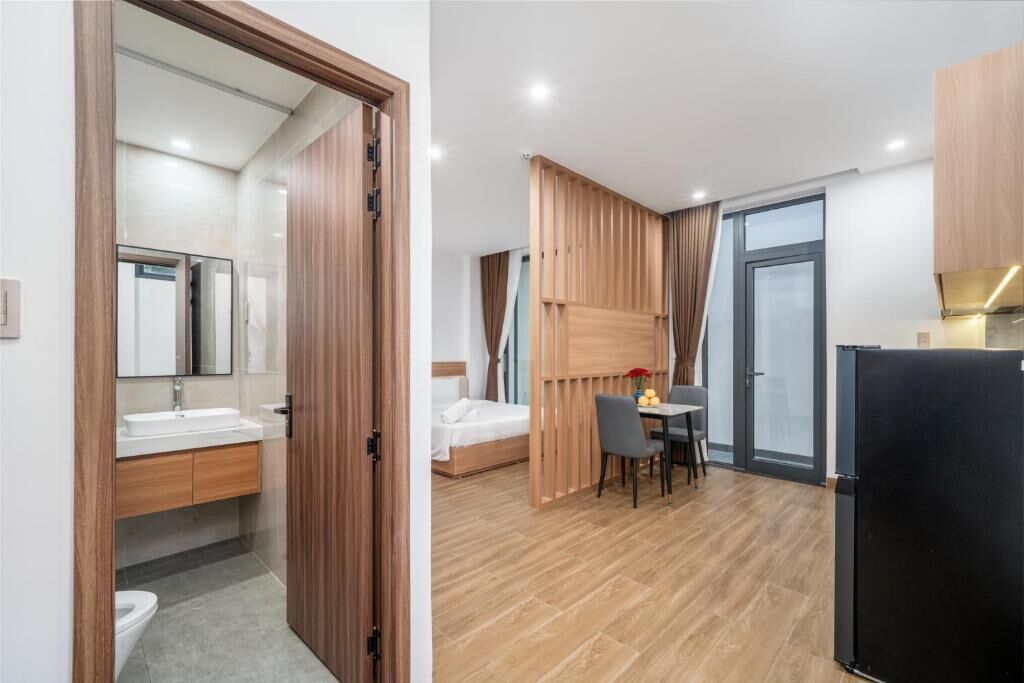 Una habitación de hotel moderna con puerta de madera, baño con espejo, cama, zona de comedor con mesa y sillas, y refrigerador.
