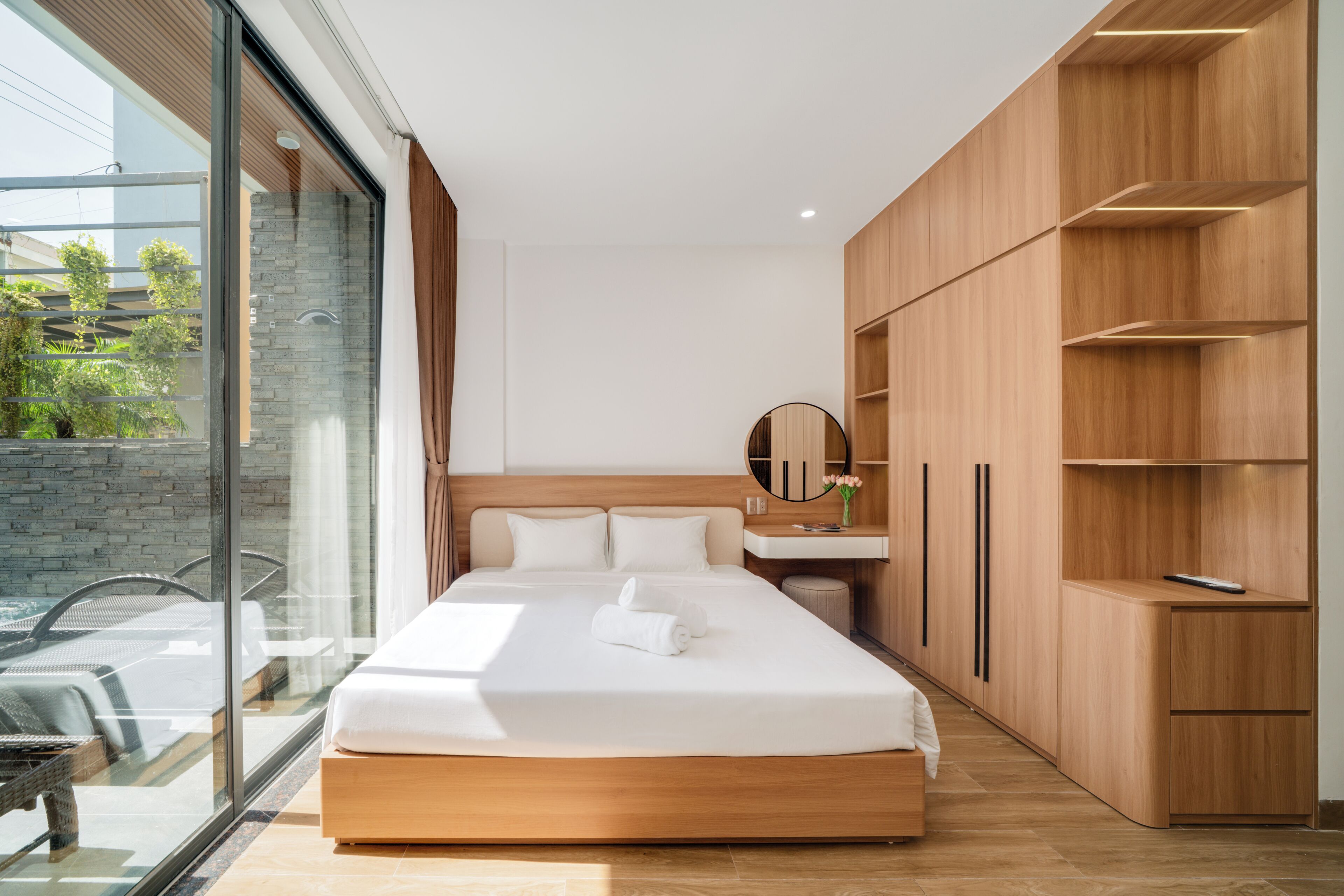 A modern hotel room with a large bed, a wooden wardrobe, a bathroom with a mirror, and a view of greenery through a glass door.