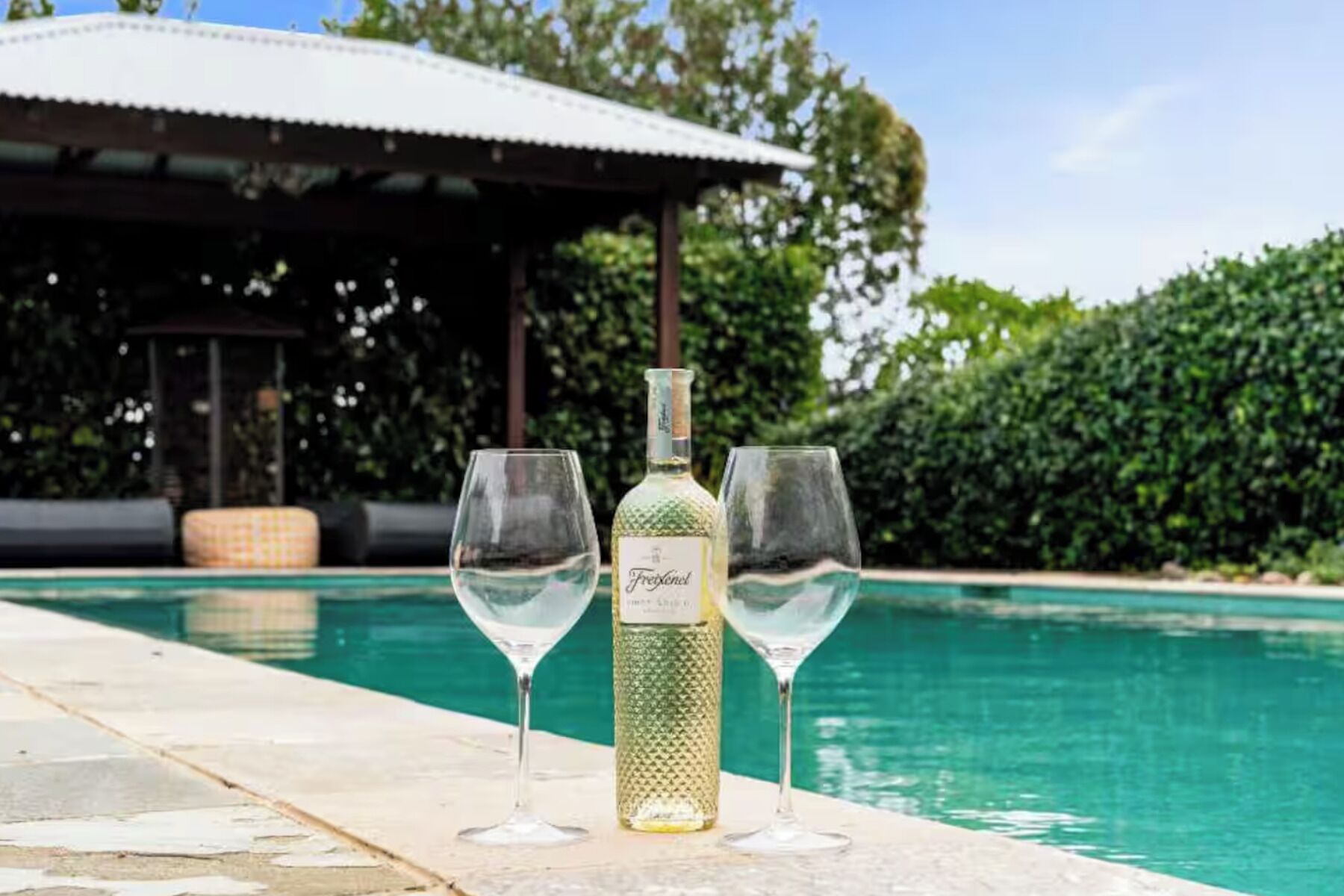 A poolside setting with a bottle of Freixenet wine and two glasses, a covered seating area, and a pool.