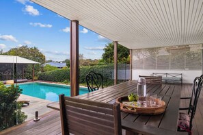 Um deck de madeira com piscina, móveis de exterior e uma área coberta com vista para a piscina e vegetação.