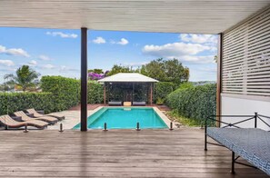 Uma área de piscina com espreguiçadeiras, um espaço de estar coberto e vista para o verde ao redor.