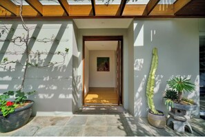Uma entrada moderna com teto de madeira, piso de pedra e diversos vasos de plantas, incluindo um cacto alto.
