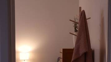 A room with a wooden floor, a bedside table with a lamp, a chair with a blanket, and a coat rack with a coat.