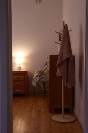 A room with a wooden floor, a bedside table with a lamp, a chair with a blanket, and a coat rack with a coat.