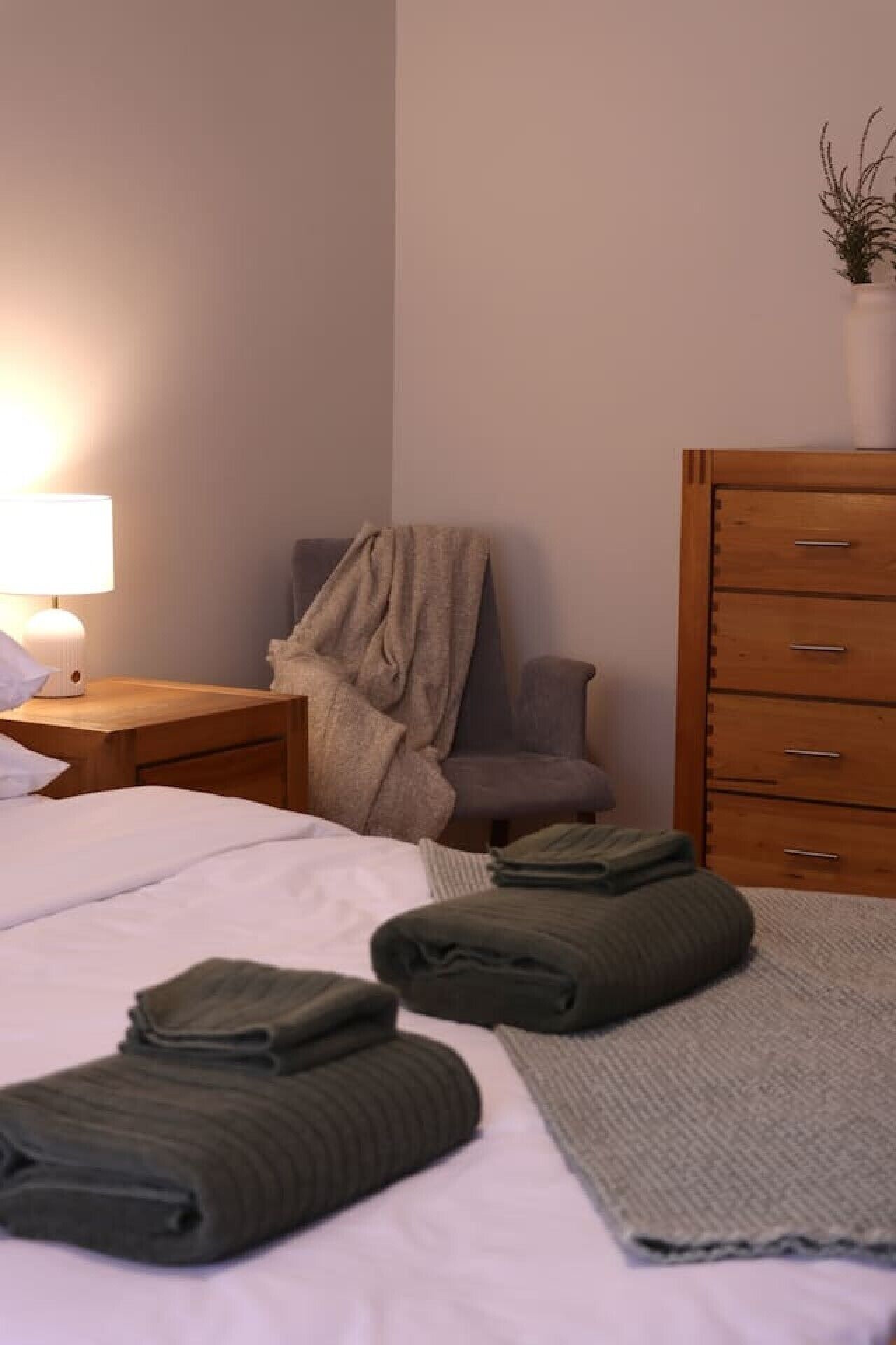A neatly made bed with folded towels, a bedside lamp, a chair, and a wooden dresser with drawers.