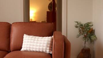 A brown leather sofa with a checkered pillow, a wooden floor, a small rug, a plant, and a door leading to another room.