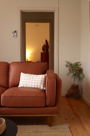 A brown leather sofa with a checkered pillow, a wooden floor, a small rug, a plant, and a door leading to another room.