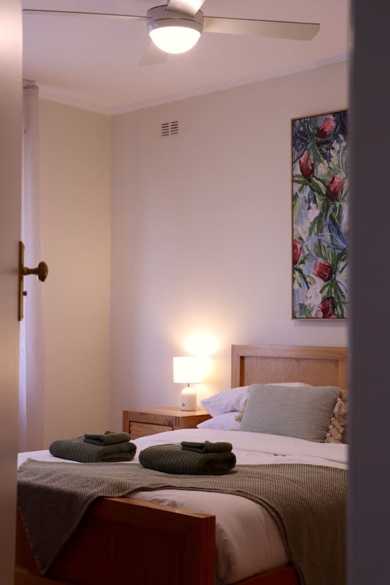 A hotel room with a bed, a bedside lamp, a painting on the wall, and a ceiling fan.