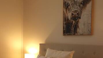 A hotel room with a large bed, two pillows, a blanket, a nightstand with a lamp, and a painting of a cow on the wall.