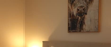 Hotellihuone, jossa on suuri sänky, kaksi tyynyä, peitto, yöpöytä lampun kanssa ja seinällä lehmäaiheinen taulu.