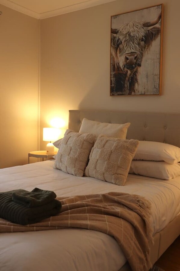 A hotel room with a large bed, two pillows, a blanket, a nightstand with a lamp, and a painting of a cow on the wall.