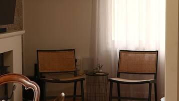 A room with a dining table, two chairs, a small round table with a vase, and a window with sheer curtains.