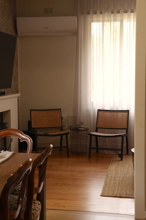 A room with a dining table, two chairs, a small round table with a vase, and a window with sheer curtains.