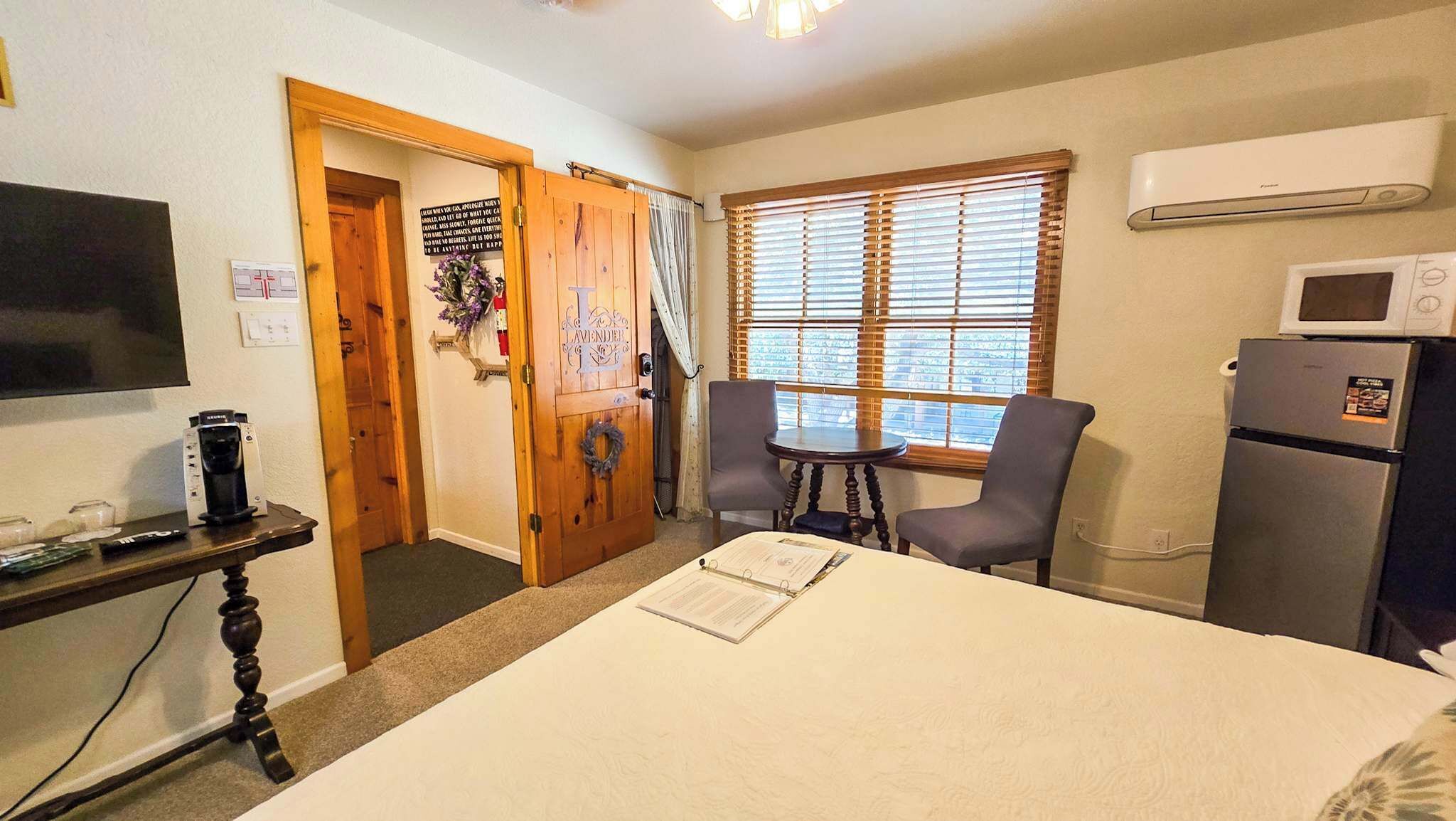 Una habitación con cama, escritorio con cafetera, silla, ventana con persianas y refrigerador.
