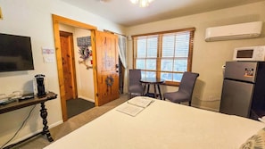 A room with a bed, a desk with a coffee maker, a chair, a window with blinds, and a refrigerator. - Lavender  Gruene Cottages (New Braunfels)
