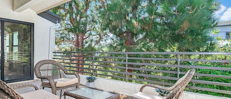 A balcony with wicker furniture, a glass table, and a view of a pine tree.