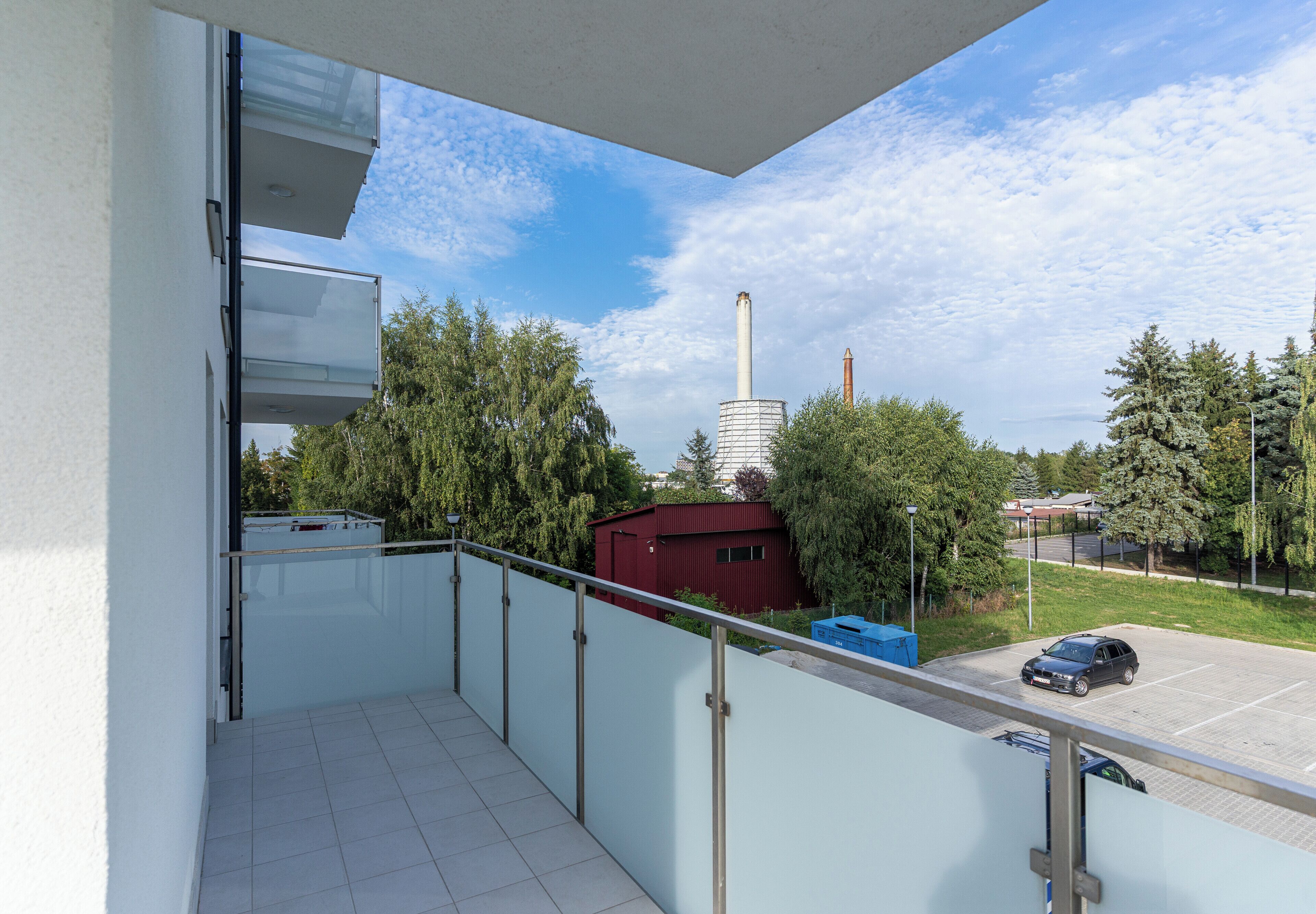 Ein Balkon mit Blick auf ein Gebäude, einen hohen Schornstein, Bäume und einen Parkplatz.