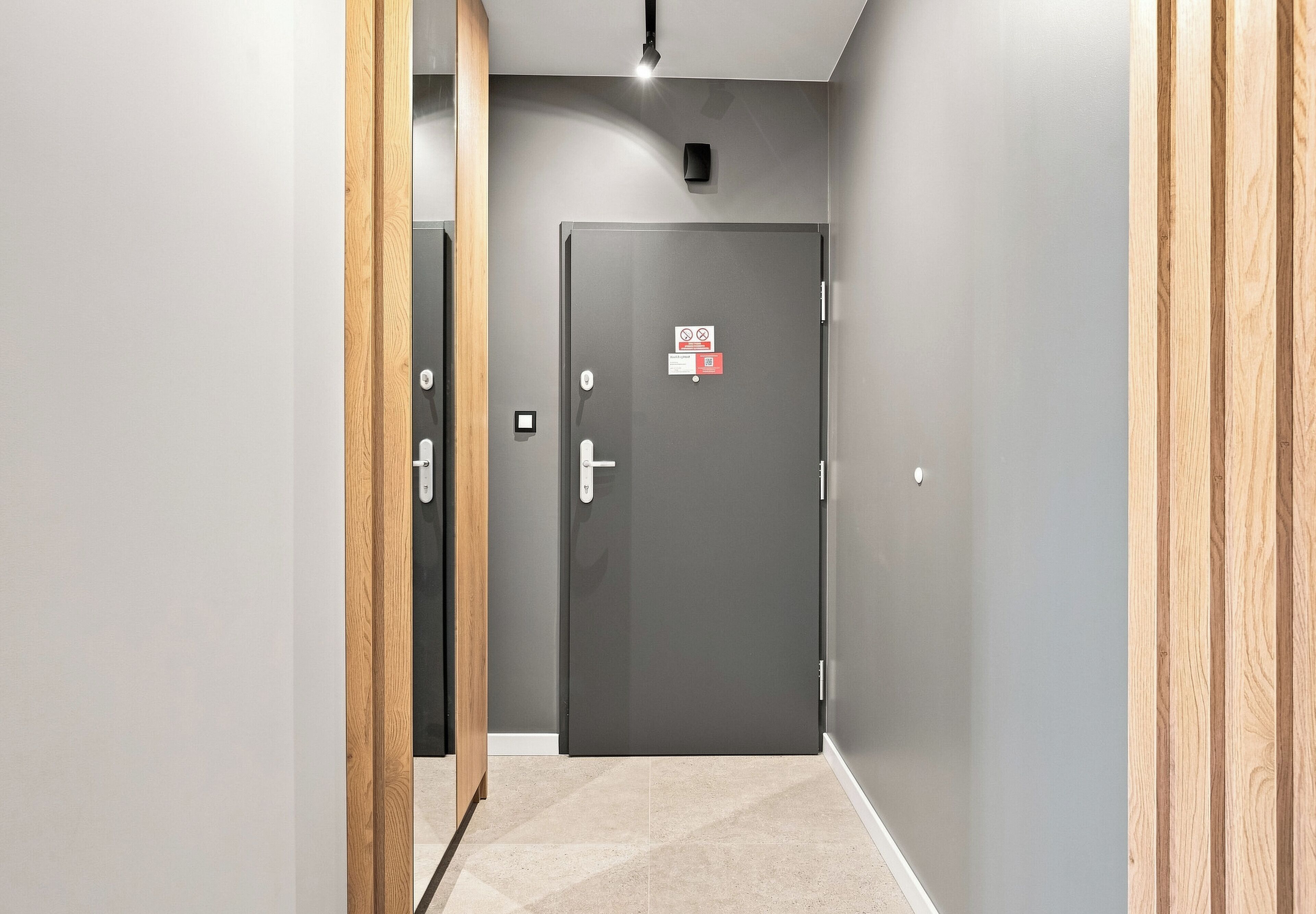 Un couloir avec une porte grise au bout, un interrupteur et une petite pancarte sur la porte.