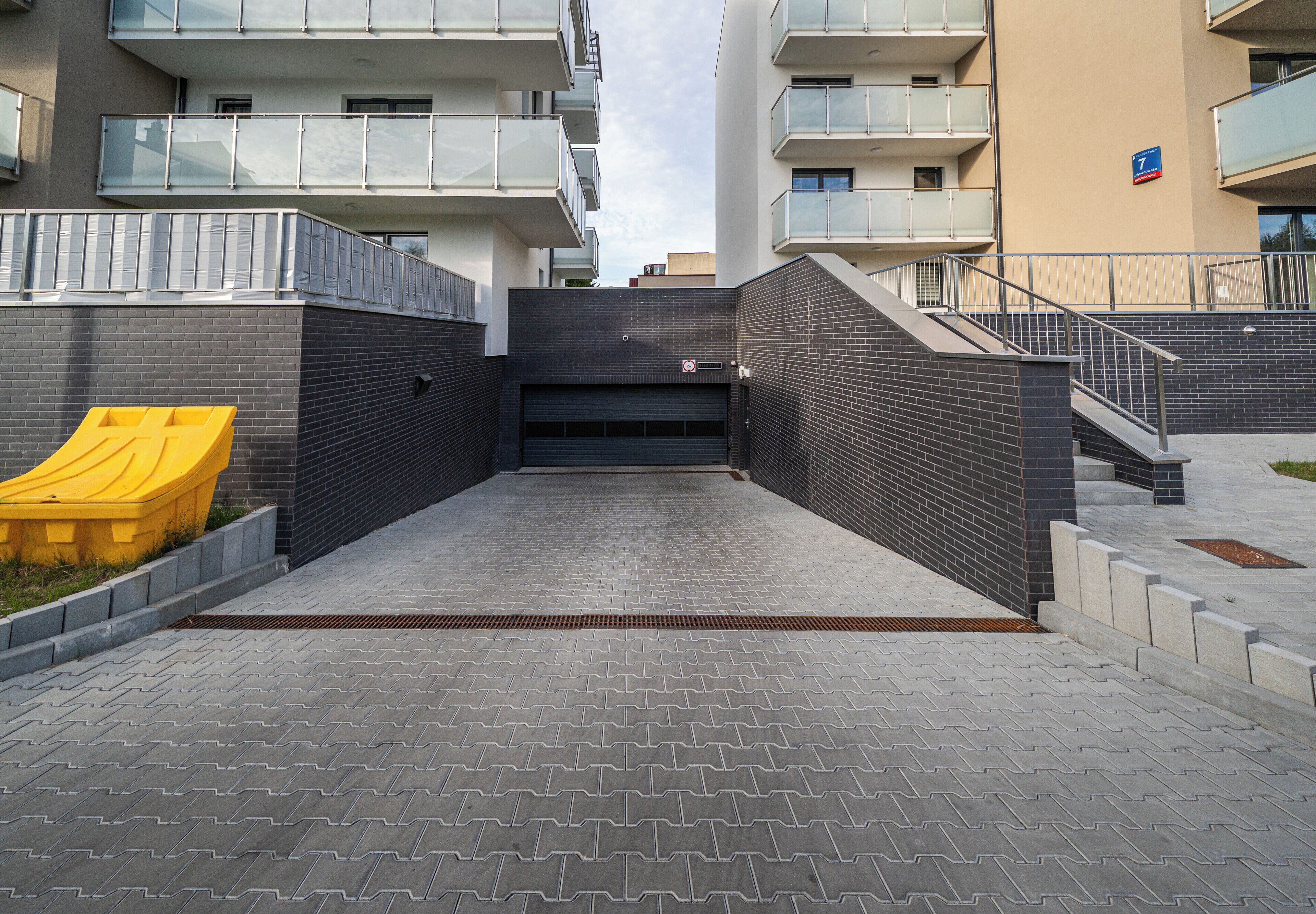 A gated entrance to a modern building with a brick facade, a yellow trash bin, and a staircase leading to an upper level.