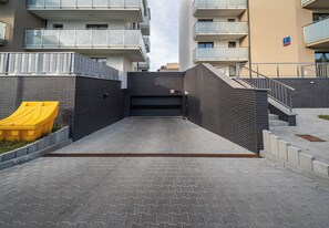 Una entrada tancada amb porta metàl·lica a un edifici modern de façana de maó, un contenidor de ferralla groc i una escala que condueix a un nivell superior.