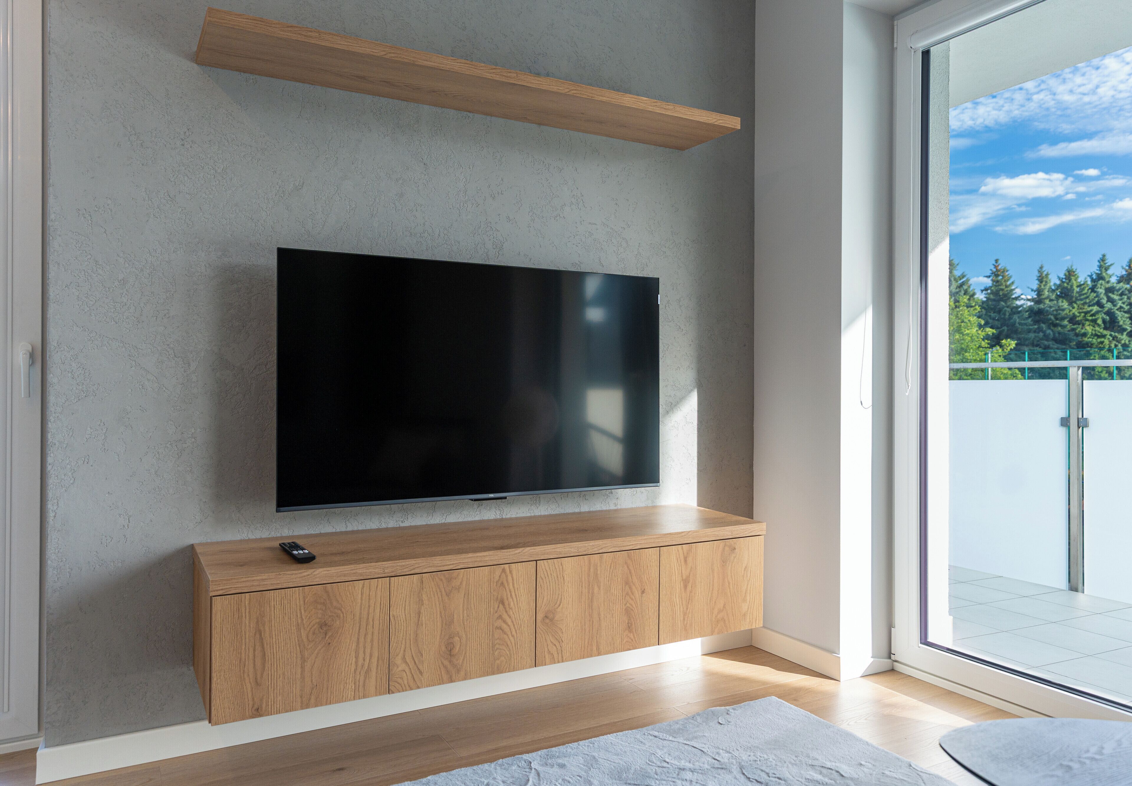 A modern living room with a flat-screen TV mounted on a textured wall, wooden cabinetry below, and a sliding glass door leading to an outdoor area.