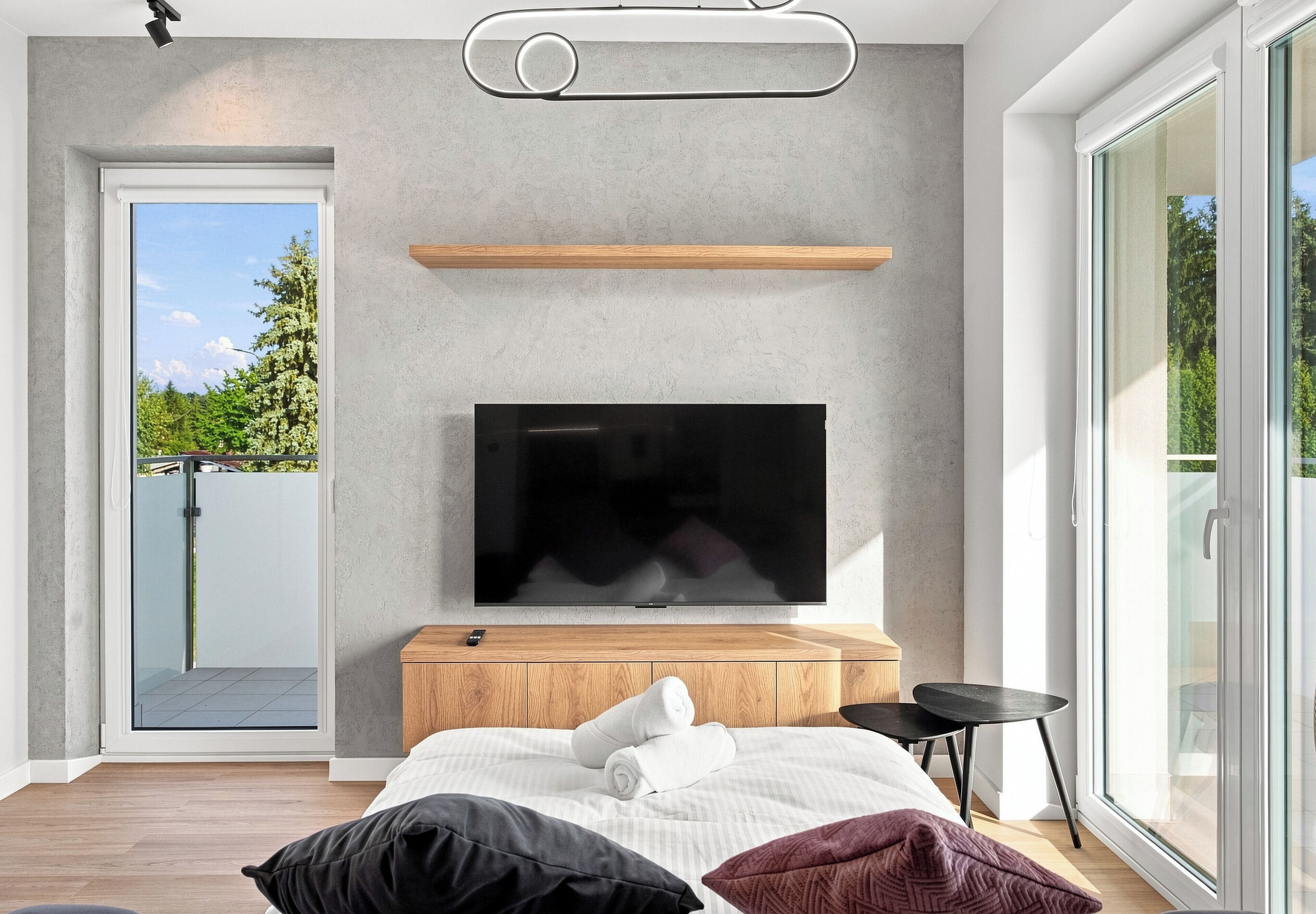 A modern bedroom with a large flat-screen TV mounted on a wooden cabinet, a glass door leading outside, and a large window providing a view of greenery.