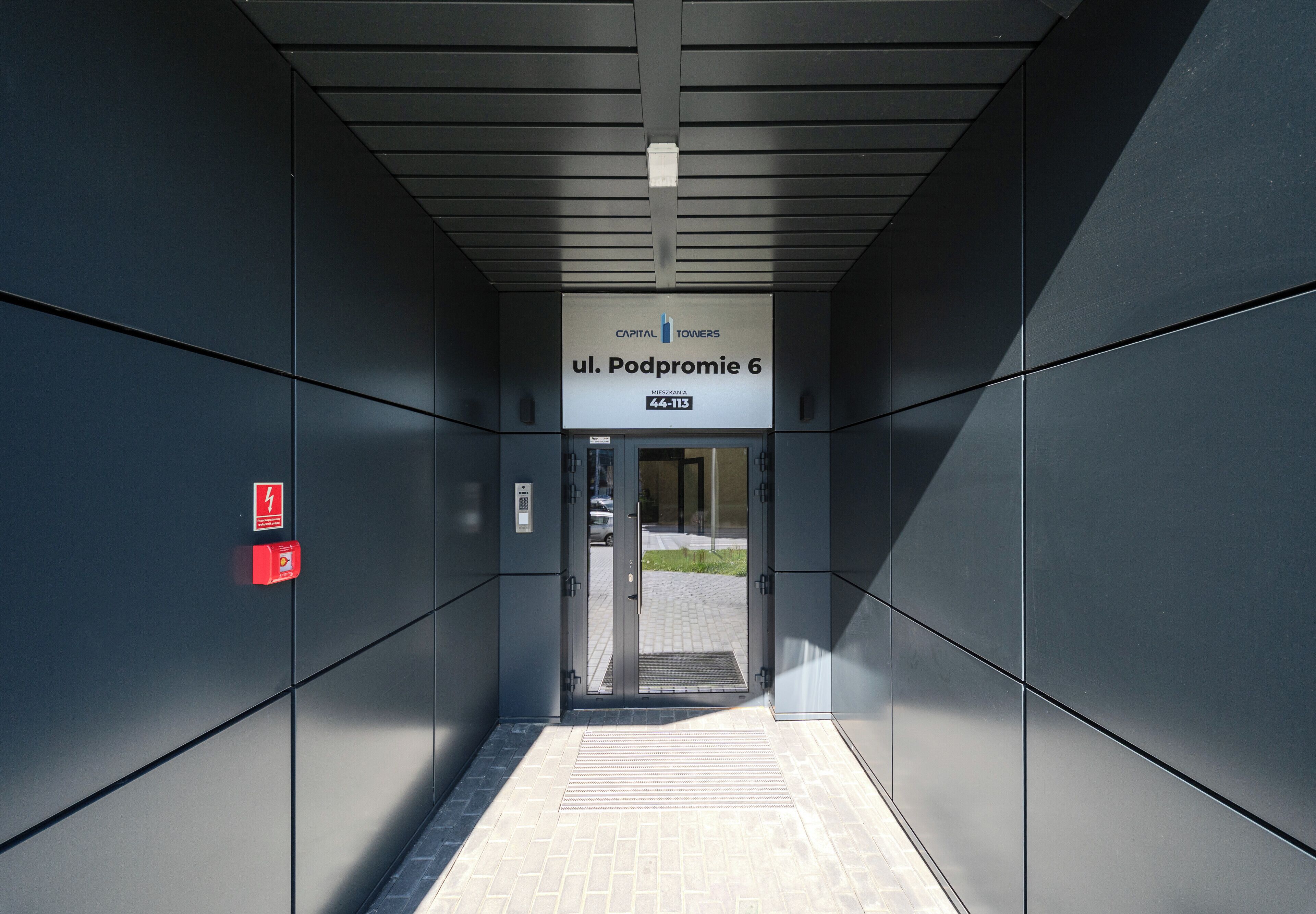 Elevatorindgang med skilt, der angiver 'ul. Podpromie 6', og en brandalarm på væggen.