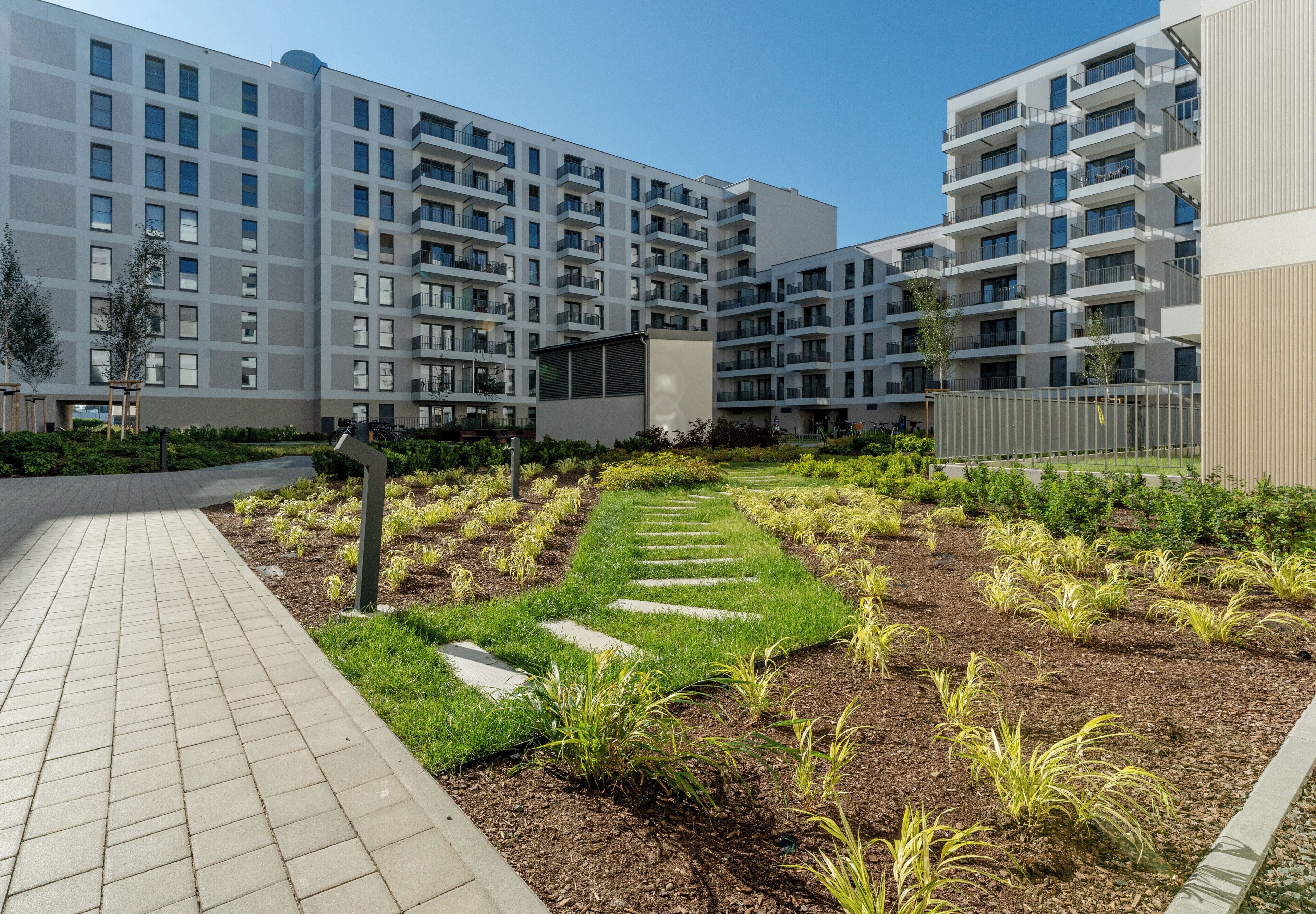 A modern apartment complex with multiple balconies, a landscaped garden area with stepping stones, and a paved walkway.