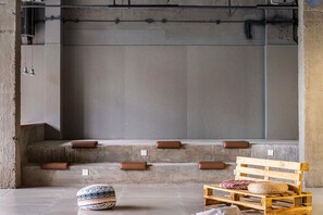 A room with a concrete wall, a bench, a patterned ottoman, and a wooden pallet with cushions.