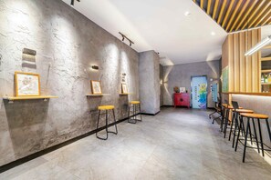 A hallway with a modern design, featuring a concrete wall with framed artwork, a ceiling with recessed lighting, and a series of bar stools along the wall.