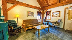 A cozy living room with a sofa, a wooden table, and a chair. There is a lamp, a framed picture on the wall, and a window with curtains. - Sweet Pecos  Gruene Cottages (New Braunfels)