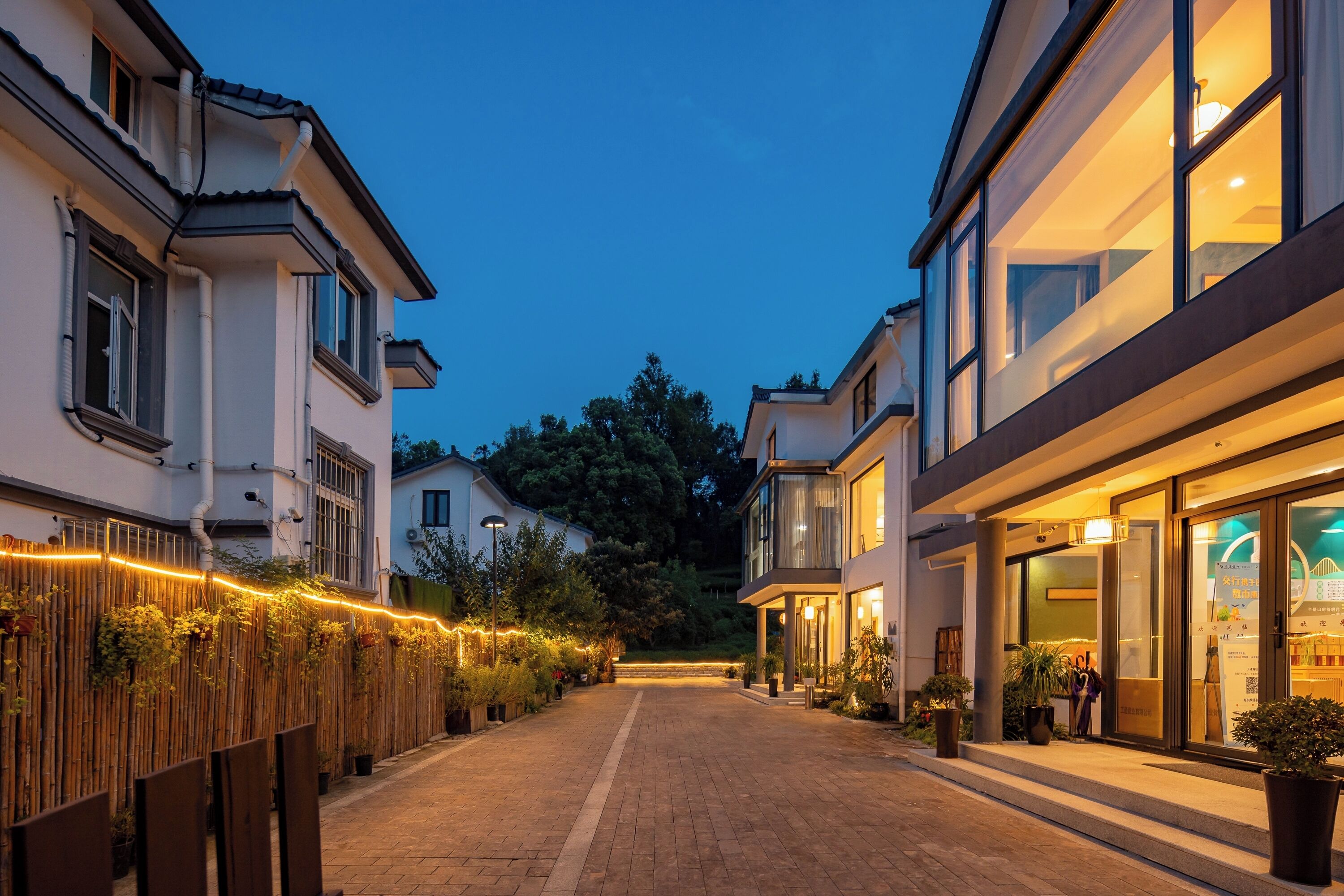 Uma rua ladeada por edifícios, com um calçadão pavimentado e uma cerca de bambu iluminada.