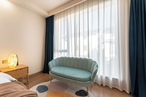A modern bedroom with a light blue sofa, a bedside table with a lamp, and a large window with sheer curtains.