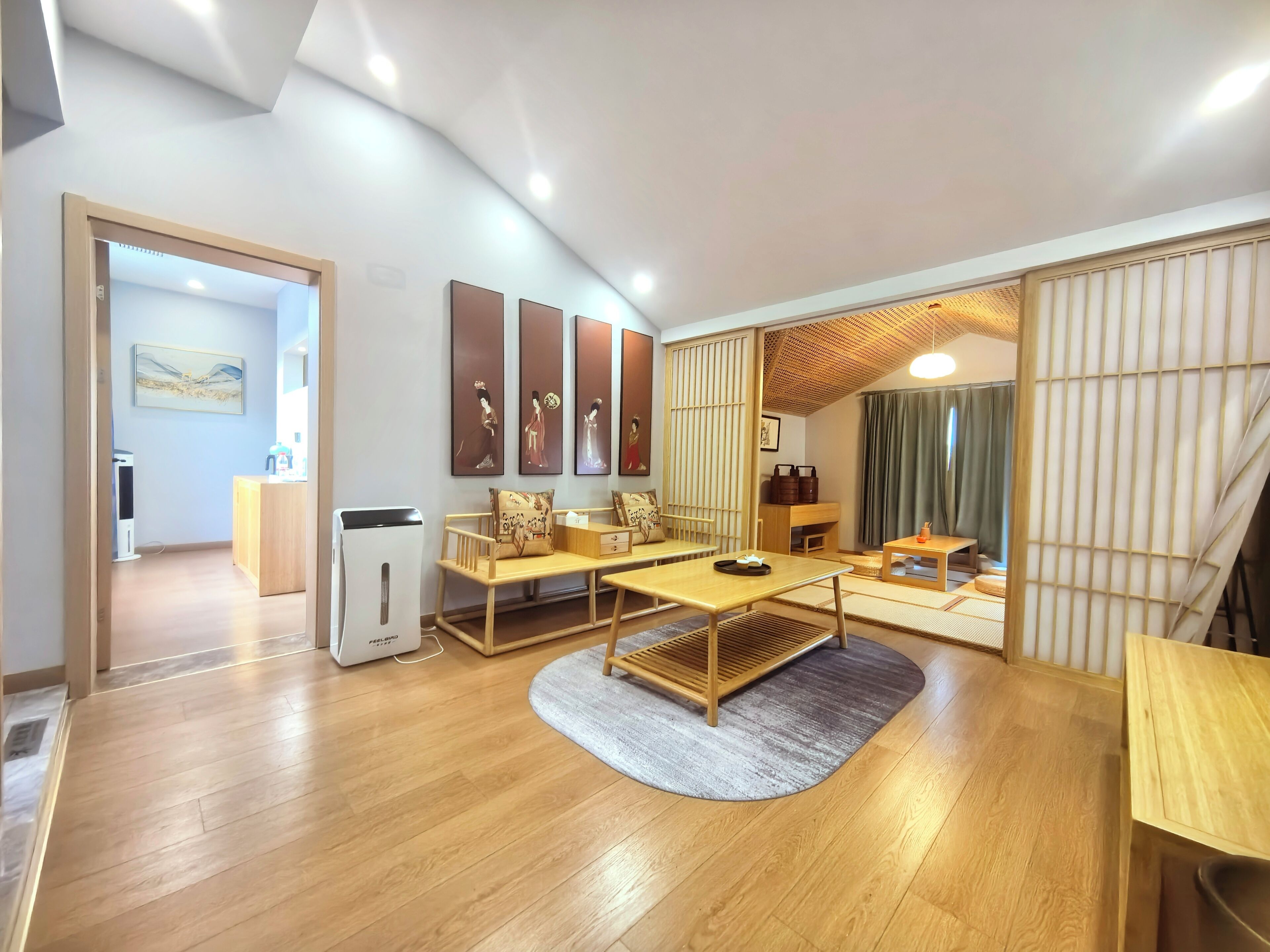 A modern living room with a wooden floor, a sofa, a coffee table, and a dining table. There are wall-mounted artworks and a sliding door leading to another room.