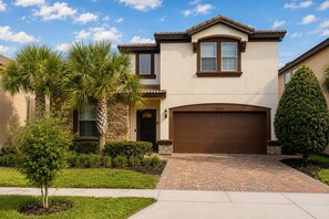 A two-story house with a brown garage door, a stone facade, and a well-maintained front yard with palm trees and shrubs. - Luxury 8BR 6BA Villa with Heated Pool, Spa, Themed Rooms, and EV Charger (Kissimmee)