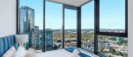 Hotellihuone, jossa on suuri sänky, sininen pääty ja näkymä kaupungin siluettiin lattiasta kattoon ulottuvien ikkunoiden kautta.