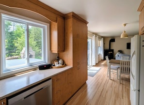 A modern kitchen with wooden cabinets, a built-in dishwasher, a dining area with a table and chairs, and a view of the outdoors through large windows. - Waterfront Cabin | Spa | Fireplaces | Gazebo (Bowman)