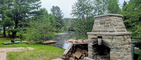 A stone-built outdoor fireplace with a fire pit, surrounded by a grassy area and a body of water.