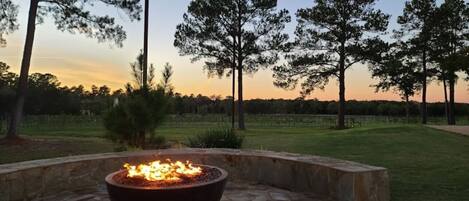 A stone fire pit with a fire, surrounded by a stone wall, in a landscaped outdoor area with trees and a grassy field in the background.
